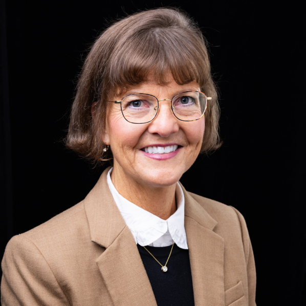 A smiling woman with short brown hair and glasses wears a tan blazer over a black top and white collar, posed against a black background.